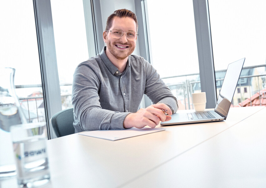 Recruiter Tammo sitzt im Konferenzraum am Tisch mit einem Laptop und lächelt freundlich.